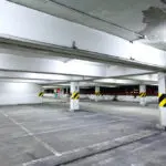 Commercial parking garage interior with deteriorating paint on the ceiling, exposed concrete surfaces, and safety-striped support columns showing signs of wear.