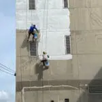 Two professional painters applying coating to a deteriorating commercial building exterior, illustrating what happens when you delay commercial painting too long.