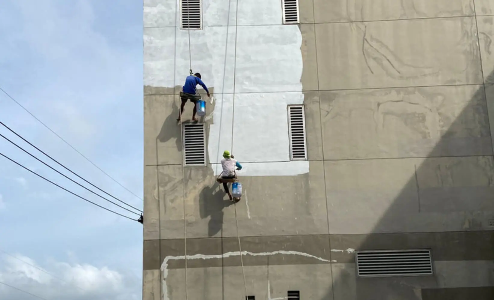 Two professional painters applying coating to a deteriorating commercial building exterior, illustrating what happens when you delay commercial painting too long.