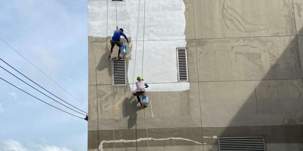 Two professional painters applying coating to a deteriorating commercial building exterior, illustrating what happens when you delay commercial painting too long.